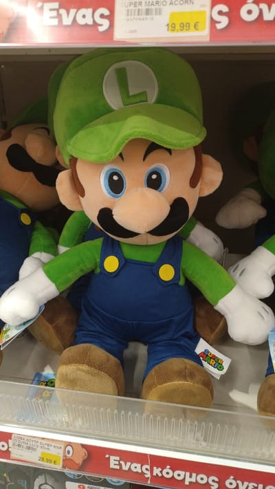 The image shows a display of Super Mario plush toys in a store, likely a toy store or department store. The central focus is a large plush toy of Luigi, recognizable by his green hat with an "L" emblem, blue overalls with yellow buttons, and green shirt. His large blue eyes, black mustache, and tan skin tone are clearly visible. To the left of Luigi, partially obscured, is a plush toy of Mario, with his red hat and blue overalls. Both toys are standing on a shelf. Above the toys, a price tag is visible, indicating "19,99 €". Further up, a red price banner displays Greek text, "Ένας" and "S όν". The background shows more merchandise on shelves and what appears to be a lighting fixture. The floor and surrounding environment are not visible, suggesting an indoor retail setting. The image does not provide information about the specific location within Pylaia Municipal Unit, Greece.