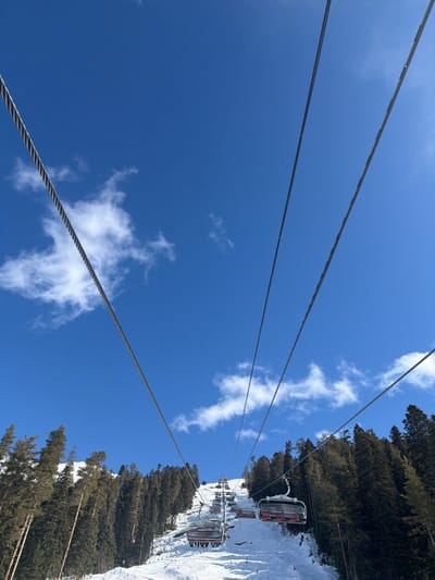 The image depicts a ski resort scene in Lunnaya Polyana, Russia. A multi-chair ski lift system ascends a snow-covered mountain slope under a clear blue sky with scattered white clouds. Several empty chairs with red seating are visible, suspended from thick steel cables that dominate the upper foreground and extend into the distance. The ski run itself is covered in packed snow, showing some tracks. Dense evergreen forests, consisting of tall coniferous trees, line both sides of the ski slope, extending upwards. No individuals are visible in the ski chairs or on the slopes.