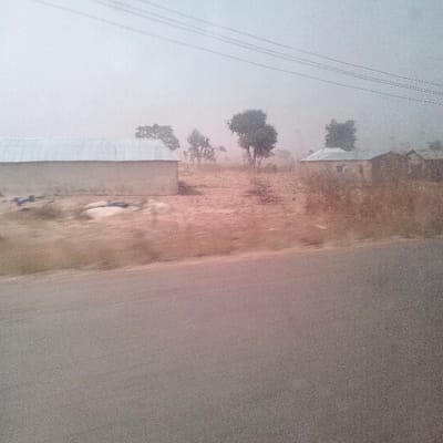The image depicts a rural or semi-urban roadside scene in Kudaru, Nigeria, under hazy or dusty atmospheric conditions. A grey asphalt road occupies the lower portion of the frame. To the left, a single-story building with light-colored walls and a silver corrugated metal roof is visible, with several indistinct objects (possibly fabric) lying on the dry, reddish-brown ground nearby. Further into the midground and right, a cluster of similar single-story structures with silver roofs and light-colored walls are present, interspersed with several trees exhibiting green foliage. The ground throughout the visible area is dry and sparsely vegetated. The sky is overcast and pale, contributing to a muted color palette and reduced visibility of distant elements. Multiple thin, dark lines, consistent with overhead utility wires, cross the upper sky. No individuals are discernible within the frame.