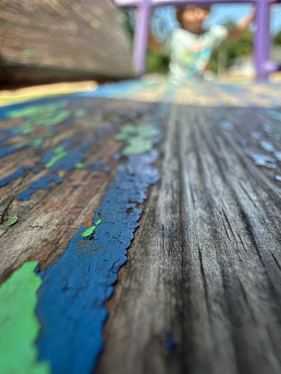 The image depicts a close-up, low-angle view of a wooden surface with peeling paint in blue and green. The wood grain is prominent, showing signs of wear. In the background, out of focus, a person appears to be on a purple structure, possibly a playground slide or swing. The setting appears to be outdoors, with greenery visible in the distance. The image does not provide specific information about its location within Piatykhatky, Ukraine.
