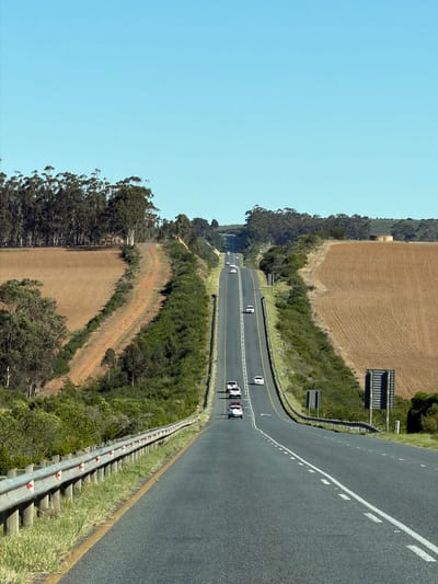 The image depicts a section of highway in George, South Africa. The road curves through a rural landscape, with brown fields and trees on either side. There are several white vehicles driving on the road. A metal guardrail runs along the edge of the highway.