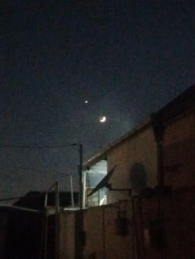 The image captures a nighttime scene in Sabunchu, Azerbaijan. In the dark, hazy sky, a crescent moon is visible, along with a small, bright point of light, possibly a star or a planet. In the foreground, a building with a tiled roof dominates the right side of the frame. A dark, shadowed fence runs across the bottom of the image. The overall atmosphere is one of low light and subdued colors, typical of a night photograph.