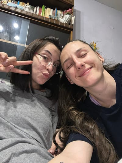 Two women are posing closely together. The woman on top is lying with her eyes closed and smiling slightly. She has brown hair and is wearing a dark blue shirt. The woman below is wearing glasses, pouting with her lips, and giving the peace sign. She has shoulder-length brown hair and is wearing a gray sweatshirt. They are in an interior setting with a bookcase visible in the background. No information to indicate location in Pamukchii, Bulgaria.