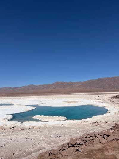 A landscape features a clear blue sky contrasting with a terrain of mountains and a unique salt flat formation. A body of turquoise water, irregularly shaped, sits amidst the cracked and textured earth, bordered by a white crust. The mountains appear barren, stretching along the horizon. The composition presents a stark, arid environment with a vibrant splash of color in the water.