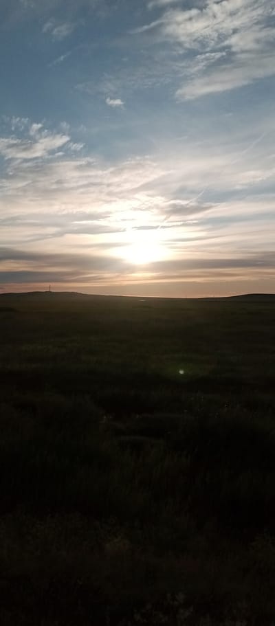 The image shows a wide, flat landscape under a cloudy sky during sunset. The sun is low on the horizon, casting a warm glow and creating streaks of light across the sky. The foreground is a dark, grassy field, with some areas appearing almost black due to the low light. In the distance, a low hill is visible, with a tall, slender structure on top, possibly a radio tower or pylon. The horizon line is sharp and distinct, separating the land from the sky. The overall impression is one of open space and natural beauty, with the sunset adding a dramatic and serene atmosphere. The location is identified as Marinovka, Kazakhstan.