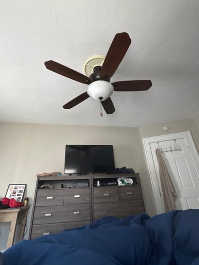 This image depicts the interior of a residential room, likely a bedroom, viewed from a low perspective, possibly from a bed. The specific address in South Bend, United States, is not discernible from the provided visual information.
The ceiling is white and features a textured, possibly popcorn, finish. A dark bronze-colored ceiling fan is centrally mounted, equipped with five dark brown wooden blades and a frosted white glass dome light fixture. A light-colored circular canopy covers the fan's electrical connection to the ceiling. A pull chain for the fan/light hangs from the fixture.
The walls are painted a uniform light beige or off-white color. A white smoke detector or carbon monoxide alarm is affixed high on the right-hand wall.
In the foreground, a dark blue comforter or blanket occupies the lower portion of the frame. No individuals are visible in the scene.
Along the main wall, a large, black flat-screen television rests atop a two-piece dark gray dresser unit. The television screen displays a reflection of the ceiling fan. The dresser unit consists of two sections; the left section features an open cubby containing a beige baseball cap and a gray fabric item, above three drawers with horizontal silver handles. The right section also has an open cubby holding a white plastic bag with green contents, above four drawers with similar silver handles.
To the left of the dresser, a light wooden bedside table is partially visible, holding a framed photograph collage, a red fabric item, and a small dark-colored bottle or container. A power cord extends downward from the table.
On the far right, a white interior door is slightly ajar. An over-the-door hanger with multiple hooks is mounted on the interior side of this door, from which a light brown or beige towel is suspended.