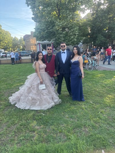 The image shows four people posing on a grassy lawn in what appears to be a celebratory outdoor setting. Two women are dressed in formal gowns, one in a navy blue dress with a beaded neckline and the other in a light pink, ruffled gown. Two men stand beside them, one in a navy suit and bow tie and the other in a maroon vest over a black shirt. Both men are wearing sunglasses.

The backdrop includes trees, a building, and a partial view of parked cars and other people in the background, including someone riding a bicycle. Based on the background context and additional information, this scene is located in the city of Hayredin, Bulgaria. The presence of formally dressed individuals suggests they may be attending or leaving a formal event, such as a graduation or prom.