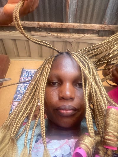 The image captures a woman receiving a hair braiding service in Ojoto, Nigeria. She is positioned centrally, looking directly forward with a neutral expression. Her dark complexion is visible, and she wears a light-colored garment with a floral or patterned design. Her hair is being styled into numerous thin, blonde box braids. Many braids are already completed and frame her face. On the right side of the frame, several finished braids are wrapped around pink hair rollers, indicating a curling step. On the left, a pair of silver scissors is held by an unseen hand, positioned near a braid, suggesting trimming. Another hand is visible at the top of the frame, manipulating a braid. A partially visible head is present on the far right. The background features a corrugated metal ceiling supported by wooden beams. A light-colored wall to the left displays several small printed images, likely examples of hairstyles.