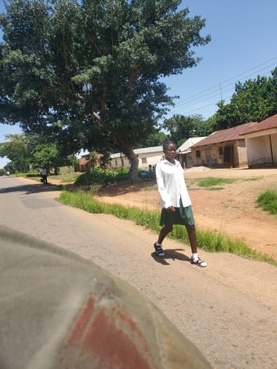 A young person stands on a paved road, facing the camera. They are wearing a white long-sleeved shirt and dark green shorts. They have black and white sandals on with white socks. The person is outdoors on a sunny day, with a clear blue sky visible. There are trees with lush green leaves on the left side of the image, and a building with a corrugated metal roof in the background. The ground on the right side of the road is dirt with some grass. The overall scene suggests a casual outdoor setting, possibly in a residential area or near a school. The time of day appears to be late morning or early afternoon, judging by the bright sunlight and the shadows cast. There is no visible text in the image.