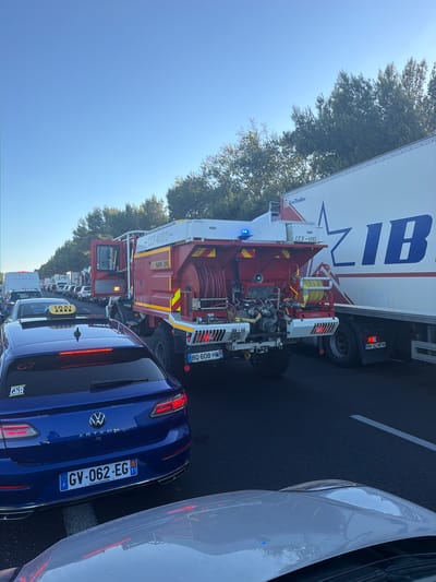 The image captures a traffic jam on a highway, likely in Villeneuve-lès-Béziers, France, indicated by the blue license plate of the blue car which features "GV-062-EG" and the European Union stars with the French flag. A red fire truck, marked "CCF 4000" and "SDIS 34," is positioned in the middle of the jam, with its door open and a driver visible inside. Next to the fire truck is a large white trailer truck with the logo "ZIB" on its side. Other vehicles, including cars and vans, are stalled behind the fire truck and trailer. A yellow taxi sign is visible on the roof of a car in the foreground. The scene is set under a clear blue sky, with trees lining the side of the highway. The overall impression is one of a halted traffic flow, possibly due to an incident involving the fire department.