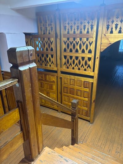 This image displays an indoor scene in Bayronovka, Russia, characterized by wooden architectural elements. In the foreground, a wooden staircase descends towards the lower right, featuring a prominent, square-topped newel post and a wooden handrail. The staircase steps and visible wooden plank flooring are painted a worn, chipped orange-brown, showing signs of significant age and use. Dominating the background is a series of light brown wooden screens or partitions. These partitions are constructed with an upper section featuring intricate, repetitive cutout patterns of ovals and wave-like forms, and a lower section composed of a grid of square raised panels. To the immediate right of these partitions, an opening or doorway leads into a darker area where the continuation of the worn wooden floor is visible. A light-colored wall is partially visible on the left side of the frame. The scene is devoid of any individuals, animals, or active subjects.