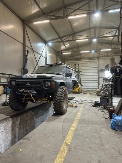 The image depicts the interior of what appears to be an automotive repair shop or garage in Musachevo, Bulgaria. A dark-colored, off-road vehicle with oversized tires is positioned on a vehicle lift. The license plate of the vehicle reads "CB 1796 PE". The shop has a high ceiling with visible metal framework and overhead lighting. Various tools, equipment, and supplies are scattered throughout the workspace. A forklift is parked in the background. A yellow safety line is painted on the floor. No people are visible in the frame.