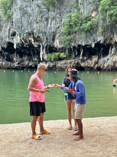 In a serene, tropical setting reminiscent of Krasom, Thailand, two individuals are captured in a tender moment. A man, with blond hair and sunglasses, stands on a sandy shore, dressed in a vibrant pink tank top and black shorts. He is holding a golden object, possibly a decorative prop. His feet are adorned with striking rainbow-colored slides. He is reaching out to a woman, who is partially obscured by his arm. She wears a blue long-sleeved shirt and light-colored shorts, complemented by a grey hat. Her arm is extended towards the man, as if in greeting or engagement. Behind them, a calm, green body of water reflects the warm glow of what appears to be late afternoon sun, with a person swimming in the distance. Towering, rugged cliffs covered in lush green vegetation form a dramatic backdrop, suggesting a natural grotto or cove. The overall atmosphere is peaceful and relaxed, hinting at a leisurely vacation or outing. The text on the man's shirt is partially visible and appears to be in a script, possibly Thai.