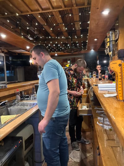 A sideways shot shows three people inside a bar or restaurant. The foreground shows a man in a light blue shirt and jeans leaning slightly over the bar. The bar appears to be wooden with a metal sink, taps, and other bar equipment. Behind him is another man standing behind the bar wearing a colorful, patterned shirt and dark pants, holding something in his hands, possibly money. Further back, another person is sitting at the bar. The bar area features shelves, bottles, and a vintage scale. The interior design includes wooden walls adorned with decorative string lights. The setting suggests the inside of a bar. This image location is in the city of Vitez, Bosnia and Herzegovina.