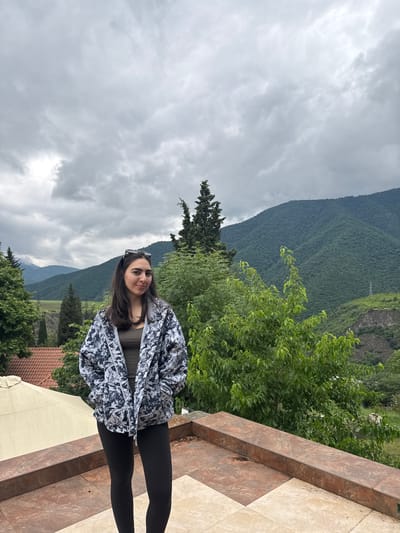 A young woman stands on a tiled patio in front of a lush green mountainous landscape. She has dark hair and is wearing a grey and white patterned jacket over a dark tank top and black leggings. Sunglasses are perched on her head. The patio has an ornate reddish-brown tiling pattern. In the background, a resort-like structure with a beige umbrella can be seen to the left, and dense green trees and hills dominate the middle ground. The sky is overcast with thick, grey clouds. A power line is visible on a distant hillside. The scene appears to be in a rural or mountainous area, possibly near Akhtala, Armenia, given the vegetation and mountainous terrain.