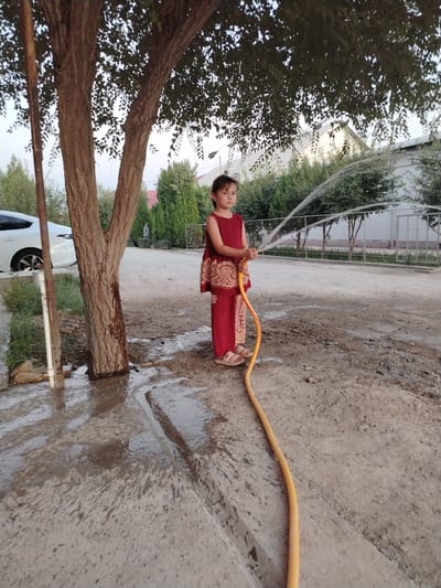 A young girl wearing a red dress with a patterned overlay stands to the right of a tree, holding a yellow garden hose from which water is spraying. She is facing the viewer, looking slightly to her left. She wears sandals on her feet. The ground around her is wet and muddy, with puddles of water visible. A white car is parked to the left of the frame, behind a tree trunk and a white pole. In the background, there are green trees and a white fence. The scene appears to be an outdoor residential area in Shovot, Uzbekistan.