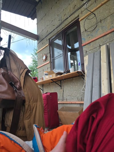 This image depicts an outdoor or semi-enclosed area adjacent to a building, observed from a low, first-person perspective. The precise location in Brilliant, Russia, cannot be determined from the visual information provided.
In the immediate foreground, at the bottom center, a human hand or arm partially clad in an orange and blue garment is visible. To the right, a significant portion of folded red fabric obscures the view. On the left, a brown leather backpack with a strap displaying the letter 'P' is suspended from a metal hook rack, alongside a crumpled brown jacket.
The midground features the exterior wall of a building, constructed from light-colored, rough-textured panels, possibly insulation material, secured by horizontally aligned metal fasteners or pins embedded in rows. Several unfinished wooden planks are affixed horizontally to this wall. A brown-framed window is present in the wall, reflecting portions of the exterior and sky. To the left of the window, a small decorative wooden clock resembling a ship's wheel is mounted. Below the window, a wooden shelf holds a hot glue gun, a roll of white paper, and a purple-topped spray bottle. Further to the left on the wall, another wooden shelf supports several cardboard boxes and a plastic container with a clear lid. Black electrical cables are routed along the wall, secured by clips, with one segment forming a loose circular coil.
On the ground below the shelves, a red wheeled suitcase is positioned to the left, and an open cardboard box is to the right. Stacked planks of wood, some unpainted and some painted grey, lean against the wall to the right of the window.
Above the main scene, on the left side, a dark brown roof soffit or overhang is visible, from which string lights with small bulbs are suspended. Below this, a light-colored ceiling or cover extends, featuring a large wooden beam that also has string lights attached. Beyond the structure, a partly cloudy sky with patches of blue is visible, with green tree foliage in the distance. No direct actions or interactions are depicted, and no other individuals are visible.