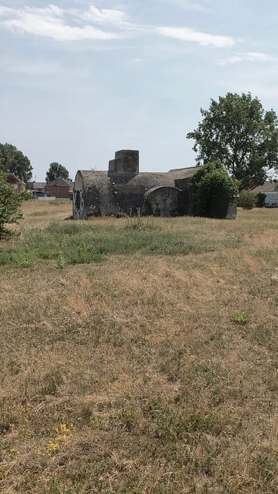 A concrete bunker, possibly a former military installation, is situated in a grassy field under a cloudy sky. The bunker appears to be in a state of disrepair, with some graffiti visible on its surface. Behind the bunker, residential buildings and trees can be seen, suggesting an urban or suburban setting. The field is covered in dry grass and some weeds. The image appears to have been taken on a bright, overcast day.