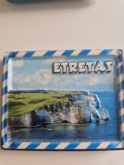 A rectangular, horizontally oriented souvenir item, likely a magnet, is depicted on a white surface. The item is located in Villeneuve-Saint-Georges, France. Its perimeter is framed by a border of alternating diagonal blue and white stripes. The central area of the item features a graphic representation of a coastal landscape. This landscape prominently displays white chalk cliffs, topped with green grass, extending into a blue sea under a light blue sky with scattered white clouds. A natural arch formation is visible extending from the cliffs into the water, and further offshore, a distinct, pointed rock formation (sea stack) rises from the water. The word "ETRETAT" is displayed in prominent, white, three-dimensional capital letters with a dark outline, centered near the top of the depicted scene. No individuals or active subjects are present in the image. The scene depicted on the item is identifiable as the coast of Etretat, France.