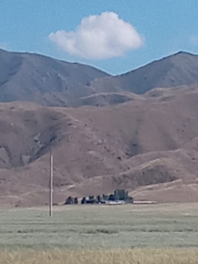 This image depicts a rural landscape with rolling hills and mountains in the background, under a light blue sky with a single white cloud. In the mid-ground, there is a cluster of structures, possibly a farm or small settlement, with a prominent pole standing in front of it. The foreground consists of a grassy field. The scene is captured on a bright day, with a shallow depth of field suggesting the focus is on the mid-ground structures. The overall impression is one of a vast, open, and sparsely populated area.