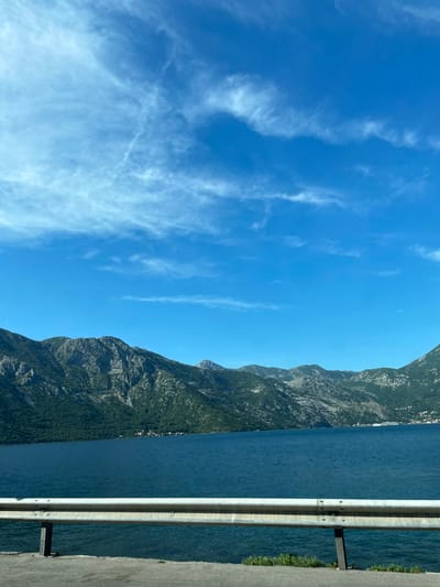 The image captures a scene in Kostanjica, Montenegro, showcasing a view of the Bay of Kotor. A portion of a roadside barrier is visible on the left. Beyond the barrier, there is a body of water, likely a part of the bay. On the opposite side of the water, a mountain range rises prominently, its slopes covered with vegetation. The sky is a clear blue with scattered cirrus clouds.