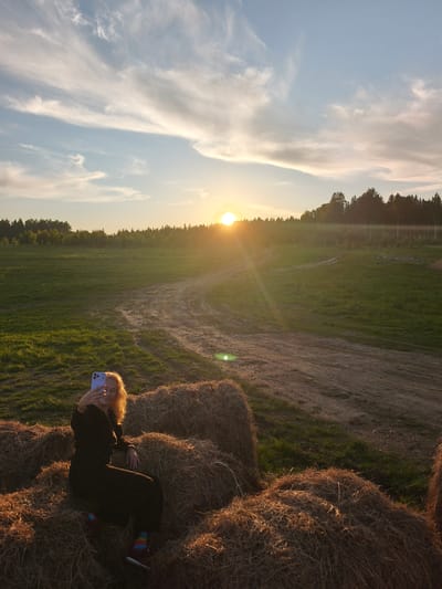 The image captures a serene rural landscape at sunset, located in Стеклянный, Russia. In the foreground, a woman with curly, light-colored hair is seated comfortably on large, stacked hay bales. She is dressed in dark, long-sleeved attire and dark pants, with visible colorful striped socks. She holds a light purple smartphone in her right hand, taking a selfie, with the device angled slightly towards her face.

The setting is an expansive open field covered in green grass, with a winding dirt path snaking through it towards the background. A dense line of trees, forming a forest, stretches across the horizon, appearing somewhat silhouetted against the sky. The sun is a prominent feature, low on the horizon on the right side of the frame, casting a bright, golden glow and warm light across the scene, indicative of either sunrise or sunset, most likely sunset given the warm hues. Wispy, elongated clouds are scattered across the light blue and orange-tinged sky. The overall atmosphere is peaceful and warm, highlighting the beauty of the natural environment during the golden hour. There is no visible text in the image.