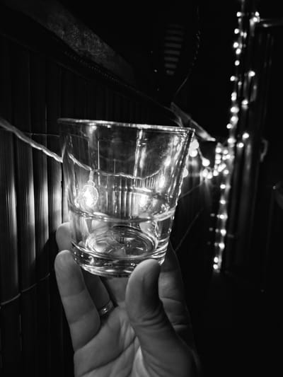 A close-up black and white shot shows a hand holding a clear glass with a few lights reflecting inside. The hand is wearing a ring. In the background, there is a striped surface and a string of blurry lights. The image is taken from a low angle, with the glass dominating the foreground. The lighting is dim, suggesting it might be evening or nighttime. The overall mood is intimate and somewhat festive due to the lights. There is no identifiable location within the city of Sinzheim, Germany.