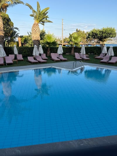 A wide shot shows a swimming pool with bright blue water and a tiled bottom. Several empty red and white striped lounge chairs with folded white umbrellas are arranged on a grassy area next to the pool. Palm trees and other lush green foliage are visible in the background, with a glimpse of the sea and distant hills under a clear blue sky. The scene appears to be a resort or hotel in the city of Chersonisos Municipal Unit, Greece.