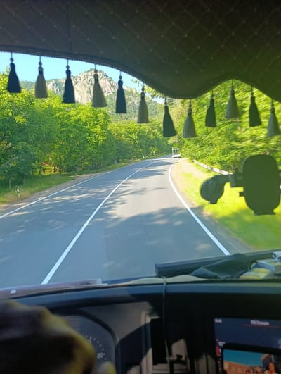 This media file captures a view from inside a vehicle, likely a truck given the dashboard details and the hand resting on the steering wheel. The foreground is dominated by the dashboard, with a hand visible on the left, appearing to be on or near the steering wheel. A digital display is visible on the right side of the dashboard, showing some text and graphics, but it is too blurry to be read clearly.
The view through the windshield reveals a paved road curving through a lush, green landscape. The road has white lane markings. On either side of the road, dense trees with vibrant green foliage line the way, suggesting a warm, sunny day with clear weather. In the distance, a rugged, rocky mountain peak rises above the trees, bathed in sunlight.
Several decorative tassels hang from the top interior of the vehicle, obscuring a small portion of the view of the sky and mountains. These tassels are dark in color, with some appearing to be black and others a dark green or gray. There is a white vehicle visible further down the road, moving away from the camera.
The overall scene depicts a scenic drive through a natural environment, likely in a mountainous region during the daytime. The sunlight suggests it is likely morning or afternoon. The image evokes a sense of travel and exploration through a beautiful, natural setting. There are no discernible emotions conveyed, and no specific event is taking place beyond the act of driving. No readable text is present in the image other than on the blurry digital display. The location context provided places this scene in Hovk, Armenia.