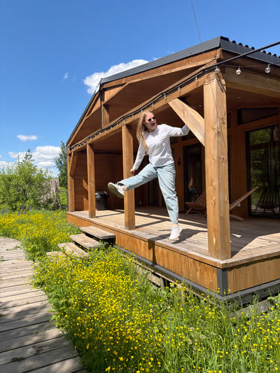A vibrant outdoor photograph captures a young woman in a playful, acrobatic pose against the backdrop of a modern wooden cabin on a sunny day in Vozhoiskii, Russia.

The central subject is a woman with long blonde hair, dressed in a white long-sleeved top, light blue jeans, and white sneakers. She wears sunglasses and has a wide, open-mouthed smile, indicating joy and playfulness, as she suspends herself horizontally, holding onto a thick wooden beam of the cabin's porch. Her body is stretched out, suggesting a moment of fun and agility.

The setting features a well-constructed wooden cabin or house with a spacious porch, characterized by robust wooden beams, posts, and flooring. A dark roof, possibly metal, is visible, along with string lights (fairy lights) attached to the eaves of the porch. A window on the cabin reflects green foliage. The ground around the cabin is covered in lush green grass dotted with numerous small yellow flowers, resembling buttercups, creating a cheerful, natural landscape. A wooden plank pathway or boardwalk runs alongside this flowering area.

The sky is a clear, brilliant blue with a few fluffy white clouds, indicating bright, sunny weather during the day, likely late morning or early afternoon, as evidenced by the distinct shadows cast by the wooden structures. There is no visible text in the image. The overall scene conveys an atmosphere of joyful outdoor leisure in a scenic, natural environment.