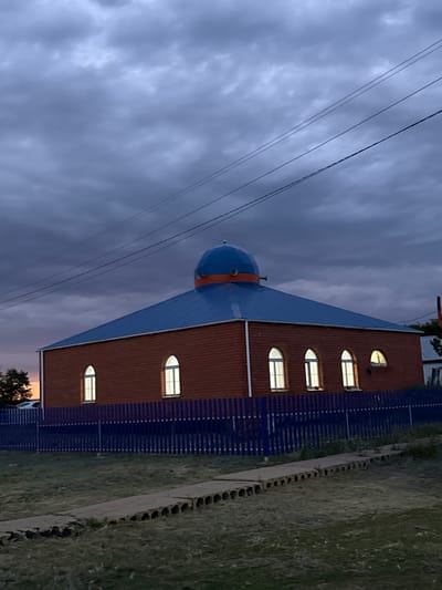 This is a vertical, eye-level, full shot of a building with a dome at dusk in a rural setting. The building has a terracotta tiled roof and a blue, domed roof with an orange band around its base. Several arched windows with internal lights are visible along the side of the building. A blue fence runs along the side of the building, and beyond that, a grassy area is visible. Power lines are strung across the cloudy sky, which is a deep purple and grey, indicating twilight. The overall mood is calm and serene, with a touch of melancholy due to the dim light and overcast sky. The location is identified as Өндіріс, Kazakhstan. No people are visible in the frame.