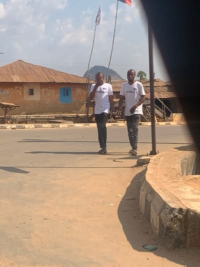 In Egbedi, Nigeria, two adult males walk on a paved road under a clear, sunlit sky. Both individuals wear identical white short-sleeved t-shirts with a dark emblem on the chest, and dark-colored trousers. The male on the left, with short dark hair and a beard, holds a dark rectangular object to his right ear, with his mouth slightly open, suggesting communication. He wears dark open-toed footwear. The male on the right, bald and clean-shaven, looks forward and slightly to the right of the viewer, wearing light-colored open-toed footwear. Long shadows are cast from their figures to the left on the road surface. In the background, a single-story building with a reddish-brown corrugated metal roof and light-brown walls is visible, featuring a prominent blue-painted window. Behind it, a conical hill rises, flanked by two tall bamboo poles, each carrying a flag; the left flag is white, the right is red and dark. The immediate foreground on the right consists of a dusty dirt shoulder bordering a concrete curb, partially obscured by a dark vertical post.