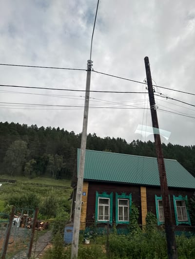 The image shows a rural scene under an overcast sky. In the foreground, a wooden house with a green metal roof and yellow siding is visible. The house has several windows with decorative turquoise trim. A wooden utility pole stands to the right of the house, and another concrete utility pole is to the left. Wires are strung between the poles. Behind the house, a dense forest of dark green trees covers a hillside. The area in front of the house appears to be a yard with overgrown vegetation, some debris, and a blue barrel. The scene is likely in a village setting, possibly in or near Uznezya, Russia, given the architectural style and the surrounding landscape. There are no people or animals visible in the image.