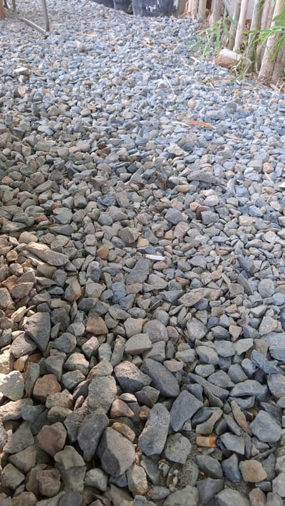 The image shows a close-up, ground-level view of a gravel path in Bireuenmeunasahdayah, Indonesia. The gravel consists of a mixture of gray, tan, and brown stones of varying sizes, some sharp and angular, others more rounded. Sunlight casts shadows across the stones, creating a textured surface. In the mid-ground, a white cigarette butt with an orange filter is visible, lying on top of the gravel. In the background, the blurry shapes of black planters or large pots can be seen, along with some thin, vertical stems and leaves, suggesting vegetation. A metallic, leg-like structure, possibly part of a chair or table, is also visible in the upper left corner. The overall impression is one of an outdoor area, possibly a garden or pathway, with some litter present.