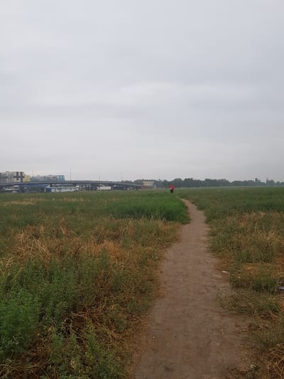 A dirt path winds through a grassy field. A person in a red shirt walks away from the camera along the path, heading towards a distant overpass and buildings. The sky is overcast. The scene appears to be on the outskirts of a city, possibly Бұлақты, Kazakhstan, with urban infrastructure visible in the distance. The foreground shows dry, brown grass and green foliage, suggesting a rural or undeveloped area within the city's vicinity.