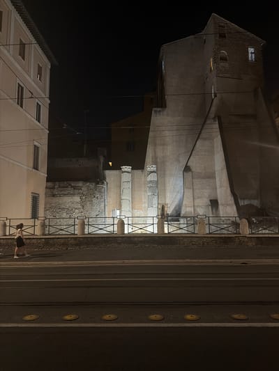 The image captures a nighttime street scene in an urban environment, possibly Rome given the architectural style. On the left, a light-colored building with multiple windows stands tall. In the center, an ancient ruin with large columns and brickwork is visible, partially obscured by a fence. To the right, a large, imposing concrete structure with a steep, angled facade dominates the background, featuring several windows, some of which are lit. A single person, a woman with dark hair, walks along the sidewalk in the foreground, dressed in a light-colored top and a dark skirt. The street in front of the sidewalk has tram tracks and yellow circular speed bumps. The sky is dark, indicating nighttime.