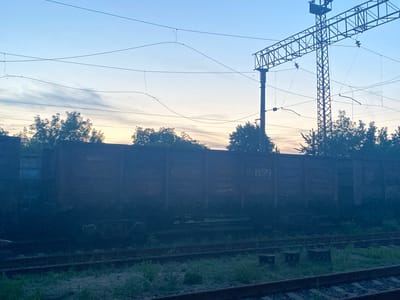 The image depicts a railway scene at dusk in Antratsyt, Ukraine. The foreground is dominated by train tracks and a long, dark freight car. The freight car appears to be a gondola or open-top wagon, made of metal, with visible markings that are difficult to decipher due to the low light and its dark color. Beyond the freight car, trees and vegetation are visible, silhouetted against the twilight sky. Overhead, electrical wires and a large metal pylon, part of the railway infrastructure, crisscross the frame. The sky transitions from a pale blue at the top to a warmer, yellowish hue near the horizon, indicating the setting sun. The overall atmosphere is serene and quiet, typical of a railway yard at the end of the day. No people are visible in the scene.