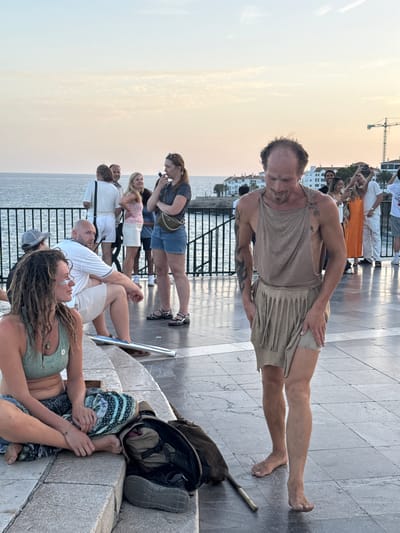 A group of people are gathered on a paved area overlooking the sea at sunset. In the foreground, a woman with dreadlocks and white face paint is sitting on a stone ledge. To her right, a man wearing a tan tunic and a fringed skirt, with tattoos on his arms, is walking and looking down. Behind them, other individuals are standing and talking. The background features white buildings, palm trees, and a construction crane. The sea is visible in the distance, with soft clouds in the sky.
