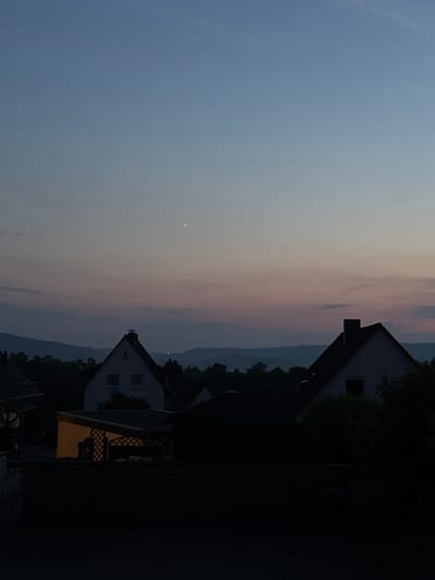 In the city of Holzminden, Germany, at dusk, a serene sky transitions from pale blue to soft pink hues. A single, bright star or planet is visible high in the sky, to the right of the image's center. Below the sky, a range of distant, shadowed hills form a dark, undulating line. In the foreground, the silhouettes of houses and trees are starkly defined against the twilight. One house, with a dark roof and lighter walls, stands out with a prominent chimney. Another structure, potentially a shed or garage, is illuminated from within by a warm, yellow light, casting a glow through a latticed opening. The foreground is dominated by deep shadows, obscuring finer details of the lower portions of the scene.