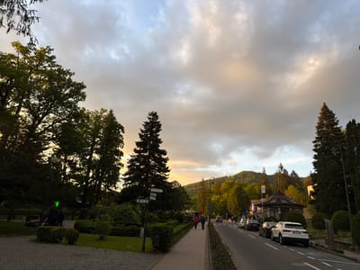 The image captures a street scene in Sovata, Romania, featuring a paved sidewalk alongside a road with parked cars. Two people walk along the sidewalk. A park area with manicured hedges and trees borders the sidewalk. A direction sign points to the right on a pole beside a tall tree. In the background, hills are visible. The sky is filled with grey and white clouds, with a touch of yellow light peeking through.