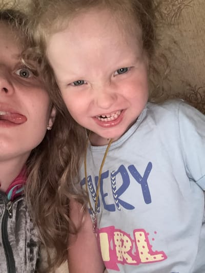 The image is a close-up selfie of two females, an adult and a child, with their faces in the foreground. The adult on the left has her tongue sticking out and is wearing a gray zipped jacket with pink and blue accents. The child on the right, with blonde curly hair, is making a grimace and wearing a light blue t-shirt with "EVERY GIRL" printed on it in blue and pink lettering. The background appears to be a textured wall or fabric in a muted brown and beige tone. The lighting is artificial and casts a warm glow. There are no identifiable landmarks or contextual clues within the image to suggest its location in Severnyy, Russia.