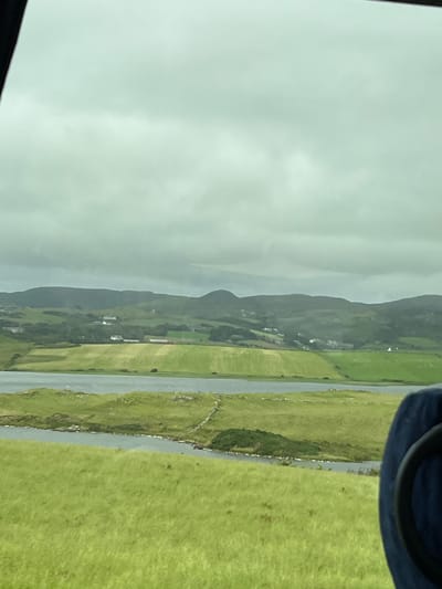 The image displays a landscape view of a rural area, likely in the vicinity of Dunfanaghy, Ireland, under a cloudy sky. In the foreground, a vibrant green grassy field stretches out. A body of water, possibly a lake or inlet, runs through the mid-ground, with grassy banks and some stone features along its edge. Beyond the water, there are more green fields, some appearing cultivated with distinct patterns. Scattered white buildings, indicative of a village or scattered dwellings, are visible on the slopes of rolling hills in the background. These hills are covered in a mix of green vegetation and darker, more rugged terrain, culminating in a hazy mountain range under the overcast sky. A dark blue object, likely a seat or part of a vehicle, occupies the lower right corner of the frame, suggesting the photograph was taken from within a moving vehicle.