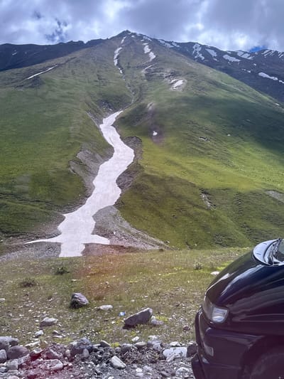 The image shows a mountainous landscape in Almasiani, Georgia. A winding strip of snow descends a grassy slope, with patches of snow also visible on the higher mountain peaks. The foreground features a rocky area with some sparse vegetation, and the front corner of a dark-colored vehicle is visible on the right side of the frame. The sky is cloudy.