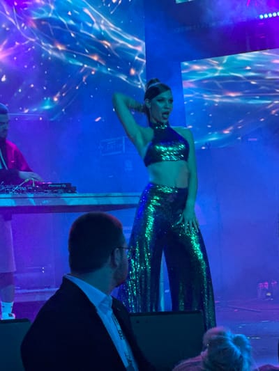 The image captures a dynamic scene at an indoor event in Tsilkani, Georgia, likely a concert or performance. In the foreground, a man wearing a dark suit and white shirt is seated, facing away from the camera and towards the stage. He appears to be an attendee.

On the stage, a female performer commands attention. She is dressed in a sparkling green sequined halter top and matching wide-leg pants, which shimmer under the stage lights. Her hair is pulled up in a high ponytail, and she is striking a confident pose with one hand behind her head and the other resting on her hip, her mouth open as if singing or speaking. The stage is bathed in vibrant blue and purple lighting, with a large, abstract digital backdrop displaying swirling patterns of light and color.

To the left of the performer, a DJ, partially visible, stands behind a DJ console. He is wearing a red jacket over a dark shirt and appears to be operating the equipment. The DJ console has a "Pioneer DJ" logo visible. The overall atmosphere is energetic and festive, indicative of a live entertainment event.