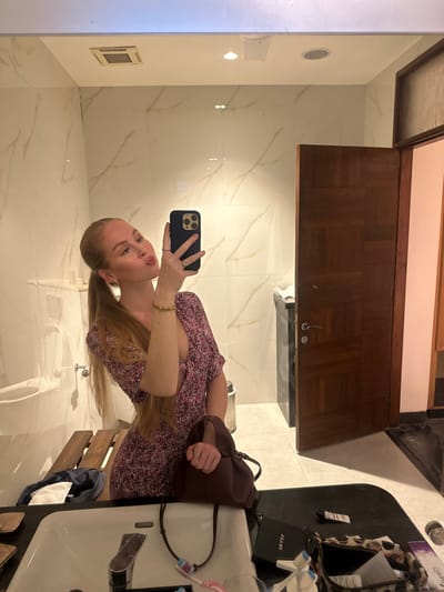 The image displays a female subject taking a mirror selfie in a restroom or bathroom located in Betalbatim, India. The subject, a fair-skinned individual with long blonde hair tied in a low ponytail, wears a short-sleeved, V-neck dress featuring a floral pattern in shades of pink and purple. She has a gold-colored bracelet on her right wrist and a ring on her left ring finger. Her right hand holds a dark-colored smartphone, directed towards the mirror to capture the reflection, while her left hand holds a dark reddish-brown handbag. Her expression shows pursed lips.

The environment includes white walls covered in marble-patterned tiles with light brown veining. A large mirror spans a significant portion of the wall. The ceiling is white, with a square ventilation grille and a circular recessed light fixture visible in the reflection. A black countertop with an integrated white rectangular sink and a chrome faucet is in the foreground. Items on the countertop include two toothbrushes (one pink, one blue), a small black rectangular case, and a leopard-print pouch containing white material. To the left, a wooden slatted bench or shelf supports a folded white cloth. To the right, a dark brown wooden door with a geometric paneled design and a chrome handle is partially visible, appearing slightly ajar and revealing a darker space beyond. A silver trash receptacle is also partially visible next to the door.