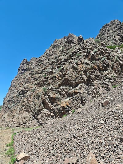 A bright, sunny day in Малиновка, Kyrgyzstan, reveals a steep, rocky mountainside under a clear blue sky. The terrain is rugged and uneven, covered with numerous loose rocks and stones of varying sizes. Sparse, dry vegetation clings to the rocky slopes, adding touches of green to the otherwise earthy tones of the mountain. A narrow, dirt path is visible along the left side of the image, winding its way up the lower part of the mountain. The overall scene depicts a natural, arid landscape with no human presence or activity evident.
