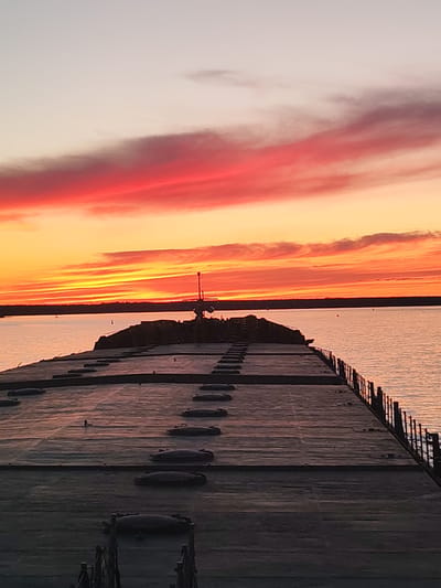 A low-angle shot reveals the flat, expansive deck of a barge at sunset. The deck is made of dark grey, rectangular panels with evenly spaced, circular access points. A railing borders the right side, separating the deck from the water. In the distance, a silhouette of land is visible against the horizon. The sky is ablaze with vibrant colors, transitioning from a fiery red to orange and yellow near the horizon, and gradually fading into a soft, light sky above. The water reflects the colors of the sunset, creating a shimmering effect. There are no people visible in the image. The scene appears to be somewhere in nature on a barge during sunset. The location in Topornya, Russia cannot be determined from the photo.