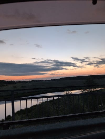 This is a photograph taken from inside a vehicle, looking out at a landscape during twilight. In the foreground, the dark metal of the car's window frame and a metal railing are visible. Beyond the railing, a body of water, likely a river or canal, reflects the sky. The water is bordered by dense green foliage and trees. The middle ground features a flat, open field or meadow, transitioning into a dark line of trees forming the horizon. The sky above is a gradient of soft blues and oranges, with scattered clouds indicating the end of the day. The scene appears to be located in or near the village of Трубичино, Russia, given the time of day and rural setting. No people or specific actions are depicted.