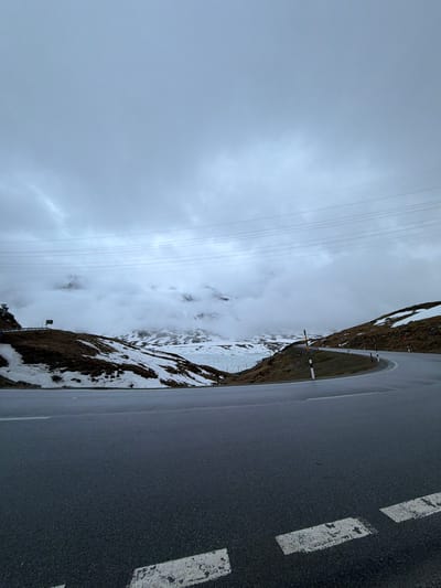 The original image is presented vertically but depicts a horizontal landscape scene of a mountainous road, likely in the Poschiavo region of Switzerland, under overcast skies.

On the right side of the frame, a dark asphalt road is prominent, appearing wet or damp, possibly from rain or snowmelt. White dashed lines mark the outer edge of the road. The left side of the image features a rugged mountain landscape. The lower slopes adjacent to the road show patches of snow interspersed with areas of dry, brown grass or sparse vegetation. Along the roadside, several slender poles or markers, some with distinctive yellow, black, and white banding, are visible, likely serving as snow poles or road markers.

Further into the midground and background, the mountains rise steeply, with higher elevations still covered in snow. Much of the mountainous terrain is enveloped in dense, low-hanging clouds and mist, partially obscuring the peaks and creating a diffused, atmospheric effect. The sky above is entirely overcast, a uniform light grey that suggests a dull or cold day. Multiple parallel power lines stretch across the upper left portion of the sky, traversing the cloudy expanse.

There are no people or specific activities visible in the scene. The overall impression is one of a remote, alpine environment, characterized by the rugged terrain, snow, and heavy cloud cover, typical of a high mountain pass during transitional seasons. No discernible text is visible in the image.