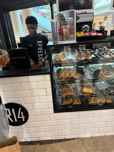 The image depicts a man wearing a black "XPRESSO" t-shirt standing behind a pastry display case in what appears to be an XPRESSO store. The display case contains various pastries, including doughnuts, muffins, and croissants, individually wrapped in plastic. The store has white tiled walls. A black circular table number sign with "R14" is visible on the side. The location is Milnerton, South Africa.